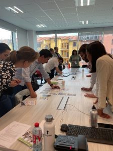 Plusieurs personnes sont debout autour d'une table. Certains se penchent sur la table pour pointer du doigts des documents et pour voir ce qu'il y a sur la table. Cette image représente des gens qui participent à une fresque.